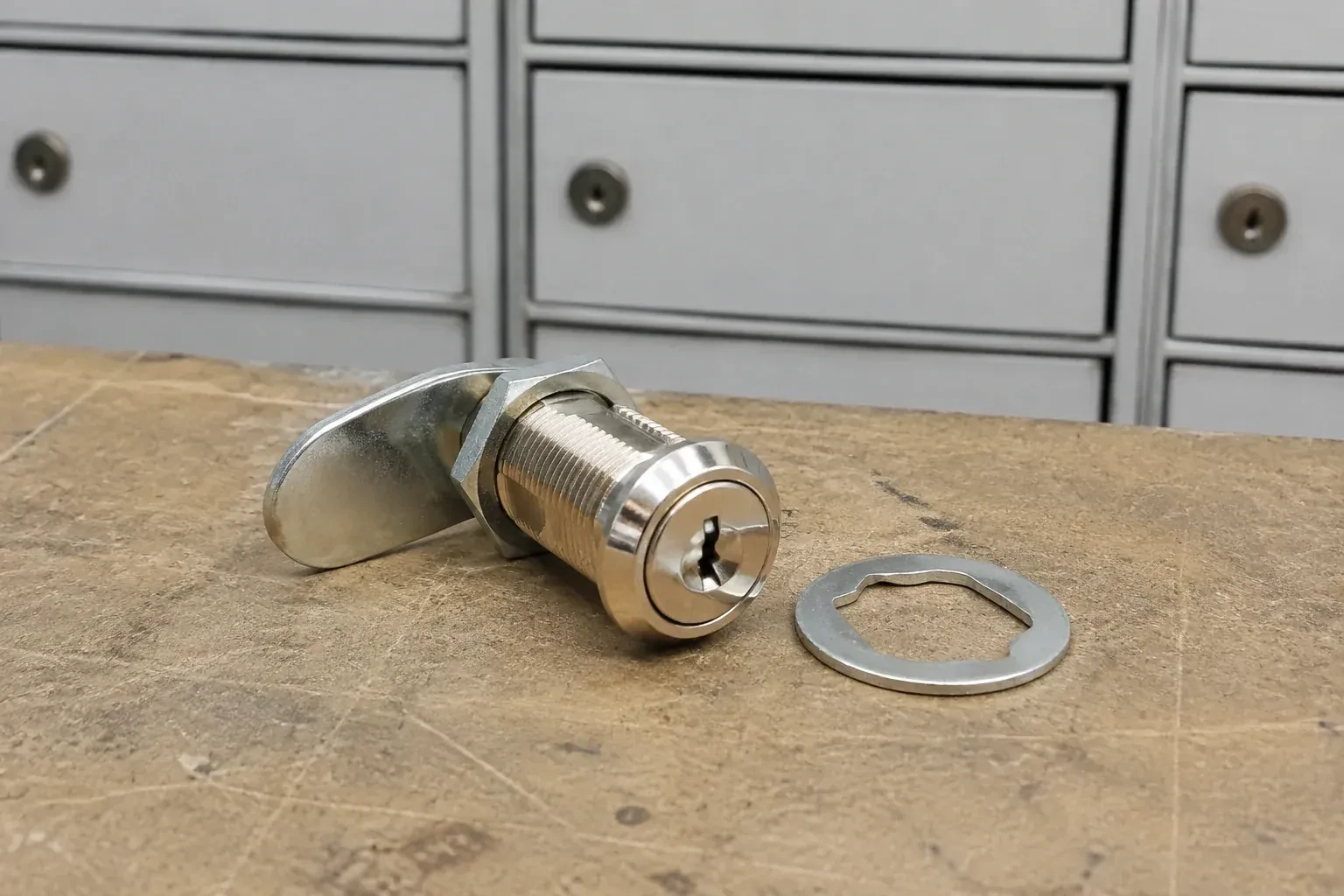 Detached threaded cam lock and mounting washer on a workbench in front of closed mail compartments. — Mailbox Lock Replacement, Clarington